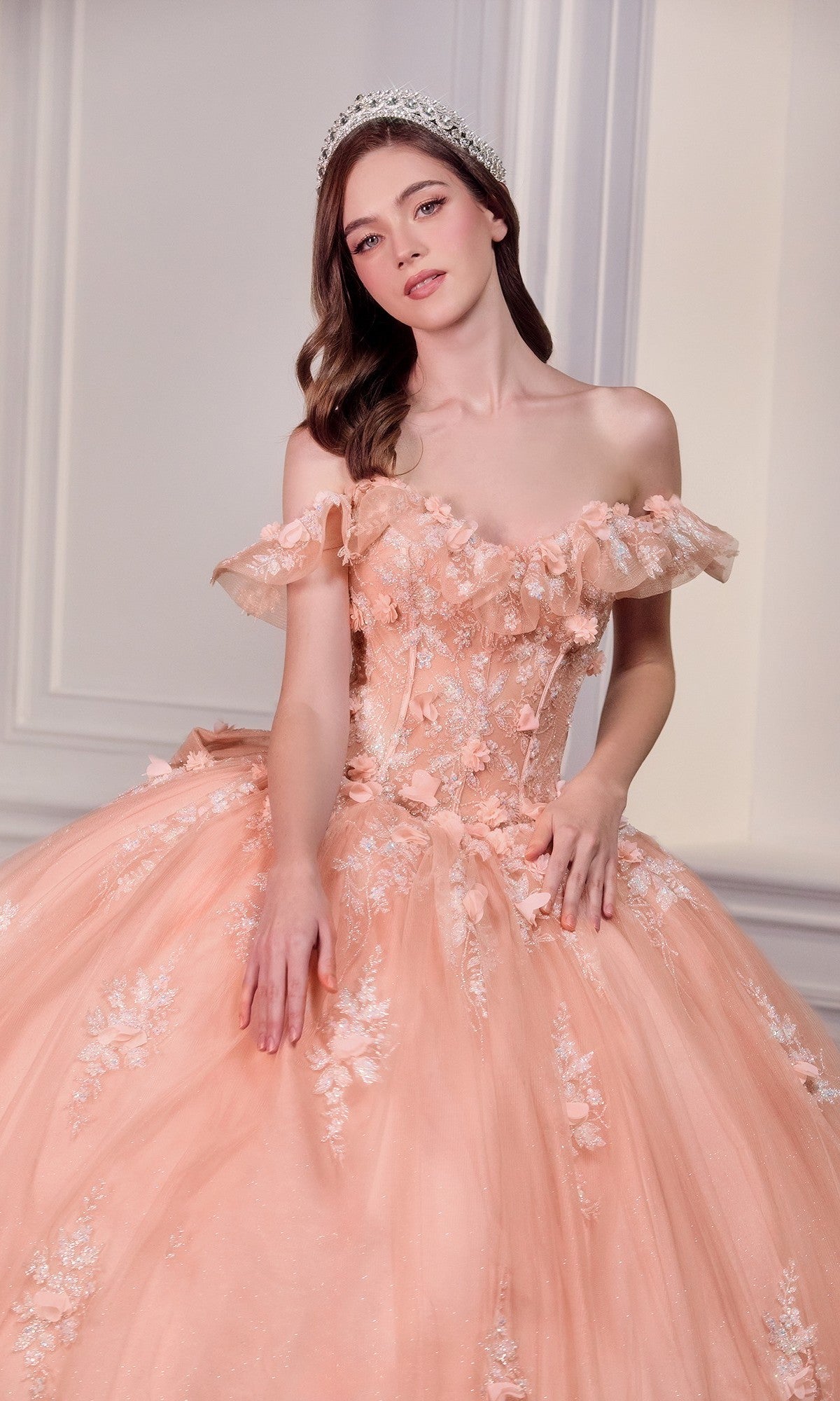 A woman poses against a light background wearing the Quinceanera Dress 15731 By Ladivine, a peach off-the-shoulder gown with 3D floral details, paired with a jeweled headband.